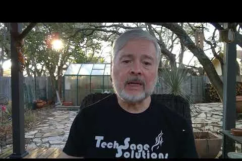 Mature man with gray hair and beard wearing Tech Sage Solutions shirt outdoors in a garden with a greenhouse behind.