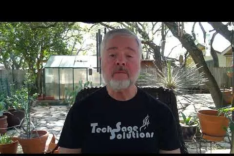 Man with gray beard wearing Tech Sage Solutions shirt sitting outdoors with plants and greenhouse in background