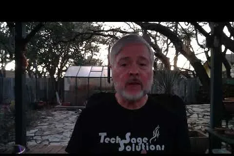 Older man with gray beard wearing a Tech Sage Solutions t-shirt sitting outdoors near a stone patio at dusk.