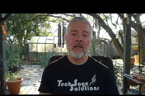 Man with gray beard wearing a Tech Sage Solutions t-shirt sitting in a backyard with plants and a greenhouse.