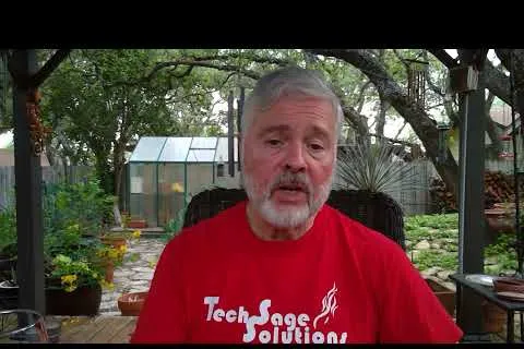 Older man with gray beard in red TechSage Solutions shirt sitting outdoors in garden with greenery and greenhouse