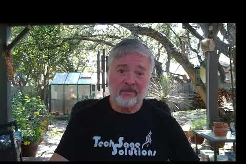 Man with gray hair and beard wearing a Tech Sage Solutions t-shirt sitting outdoors with greenery and a greenhouse behind him