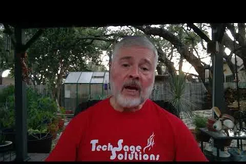 Middle-aged man with gray beard wearing red Tech Sage Solutions shirt, speaking outdoors in garden setting.