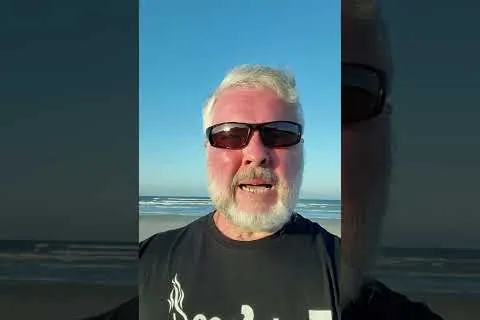Middle-aged man with white beard and sunglasses wearing a black tech tees t-shirt standing on a beach.