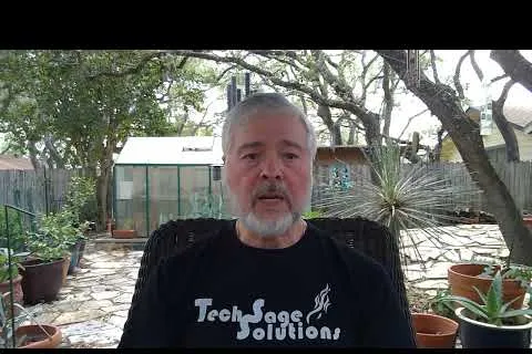 Middle-aged man with gray beard wearing Tech Sage Solutions shirt in a backyard garden with potted plants and trees.