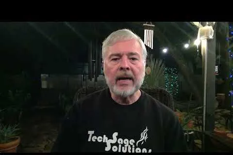Older man with white hair and beard wearing a Tech Sage Solutions shirt in a dimly lit outdoor setting.