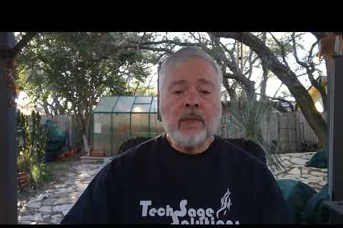 Middle-aged man with gray beard wearing a Tech Sage Solutions shirt sits outdoors in a backyard with trees and a greenhouse.
