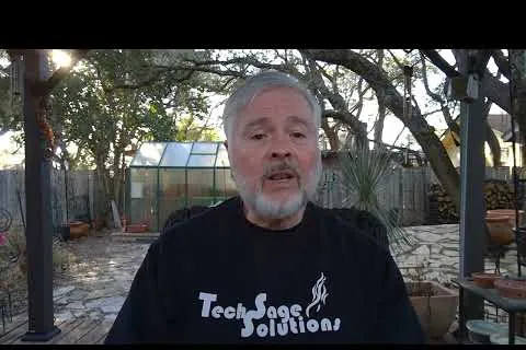 Middle-aged man with gray hair and beard wearing Tech Sage Solutions shirt speaking outdoors in a backyard with trees and greenhouse.