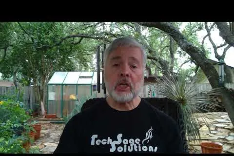 Middle-aged man with gray beard wearing Tech Sage Solutions shirt sitting outdoors near greenhouse and trees.