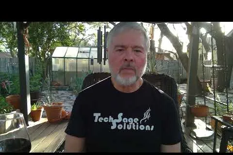 Bearded man wearing a Tech Sage Solutions shirt sitting in a sunlit garden with plants and a greenhouse.