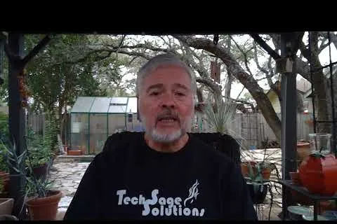 Middle-aged man with gray hair and beard wearing a Tech Sage Solutions shirt sitting outdoors with trees and plants.