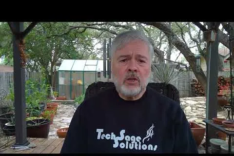 Older man with gray hair and beard wears Tech Sage Solutions shirt sitting outdoors in a covered garden patio.