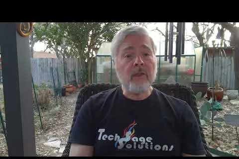Older man with gray hair and beard sitting outdoors wearing a TechAge Solutions shirt in a garden setting.
