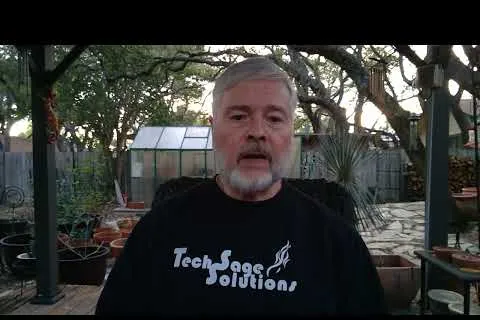 Man with gray beard wearing Tech Sage Solutions shirt sitting outdoors with plants and greenhouse in background