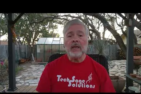 Mature man with gray hair and beard wearing red TechSage Solutions shirt outdoors with greenery and stone path