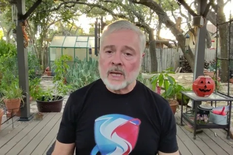 Older man with gray beard wearing a black t-shirt standing on a wooden deck surrounded by potted plants and Halloween decor.