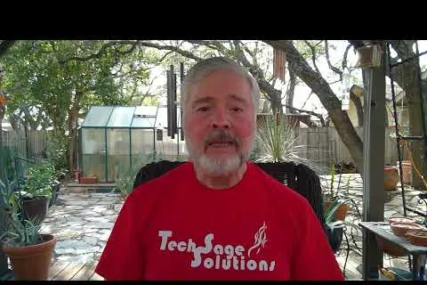 Man with gray beard wearing a red Tech Sage Solutions shirt sitting outdoors in a garden with potted plants and greenhouse.