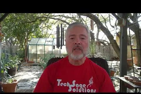 Older man with gray beard wearing red Tech Sage Solutions shirt sitting outdoors in a sunlit garden with trees and plants.
