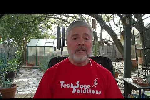 Man in a red Tech Sage Solutions t-shirt speaking outdoors with plants and a greenhouse in the background.