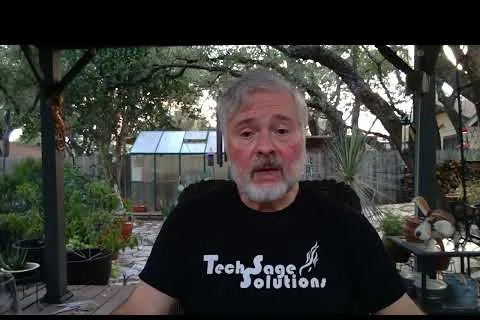 Man with gray hair and beard wearing Tech Sage Solutions shirt sitting outdoors with garden and greenhouse in background