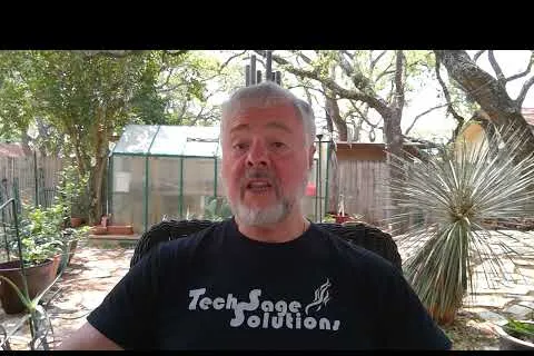 Man with gray beard wearing a black Tech Sage Solutions shirt sitting outdoors with plants and greenhouse in background