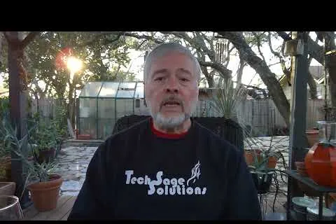 Man with gray beard wearing Tech Sage Solutions sweatshirt sitting outdoors near plants and greenhouse at sunset