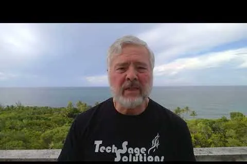 Older man with gray beard wearing a Tech Sage Solutions shirt with ocean and greenery in the background