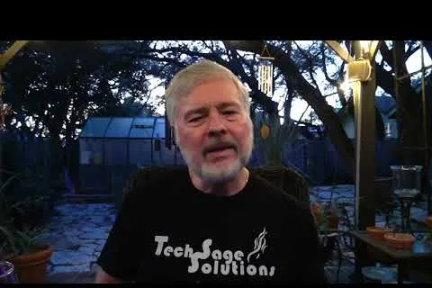 Older man with gray hair and beard wearing a Tech Sage Solutions shirt sitting outside in a backyard at dusk.