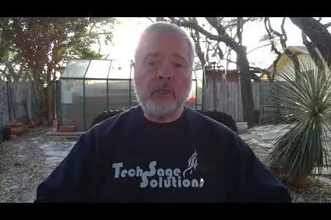 Older man with gray beard wearing a Tech Sage Solutions shirt sitting outdoors with trees and greenhouse behind.