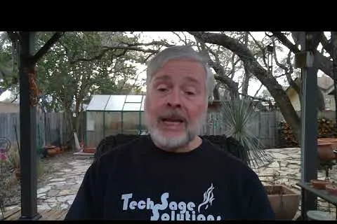 Older man with gray hair and beard wearing a Tech Sage Solutions shirt outdoors with trees and greenhouse behind him.