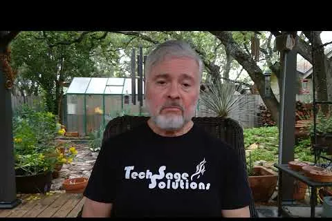 Older man with gray hair and beard wearing a TechSage Solutions shirt outdoors in a garden patio setting.
