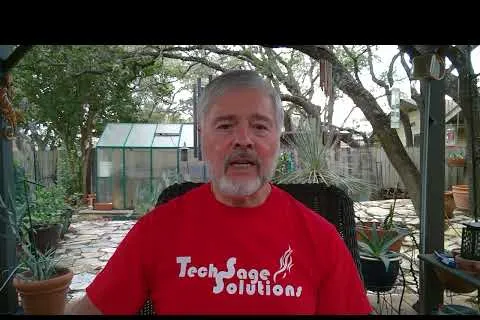Man with gray beard wearing a red Tech Sage Solutions t-shirt seated in a backyard garden with plants and greenhouse.