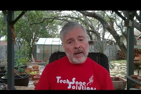 Man with gray beard wearing a red TechSage Solutions shirt sitting outdoors near plants under a wooden pergola.