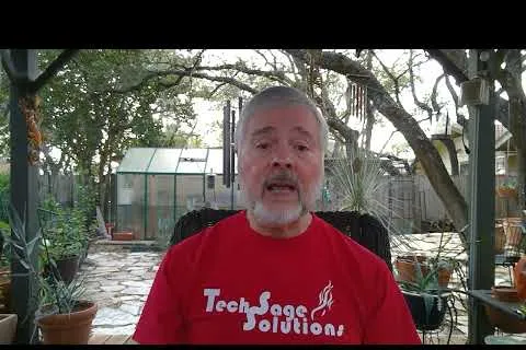 Man with gray hair and beard wearing a red TechSage Solutions shirt sitting in a backyard garden patio.