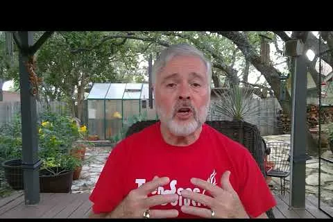 Older man with gray hair and beard speaking outdoors wearing a red shirt, surrounded by plants and a greenhouse.