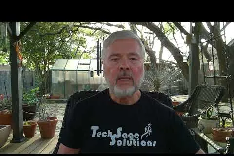 Man with gray hair and beard wearing a Tech Sage Solutions t-shirt sitting outdoors with plants and greenhouse