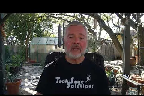 Man with gray hair and beard wearing black Tech Sage Solutions shirt sitting outdoors on a patio with plants and a greenhouse.