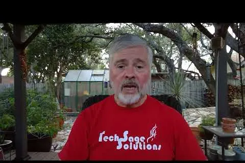Man with gray hair and beard wearing a red Tech Sage Solutions shirt in a garden with trees and plants.