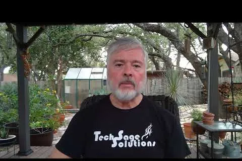 Older man with gray hair and beard wearing TechSage Solutions t-shirt sitting outdoors under a pergola with plants.