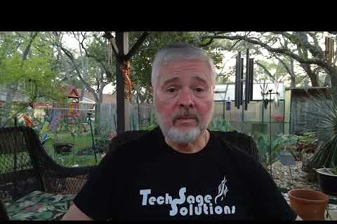 Older man with gray beard wearing a Tech Sage Solutions shirt sitting outdoors in a garden patio.