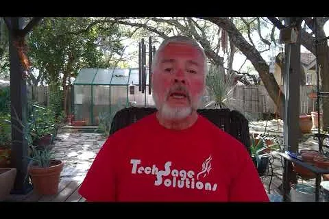Older man with gray beard wearing a red Tech Sage Solutions shirt sitting outdoors with plants and greenhouse behind him.