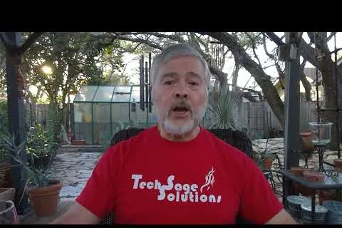 Man with gray beard wearing red Tech Sage Solutions T-shirt sitting outdoors with garden and greenhouse in background.