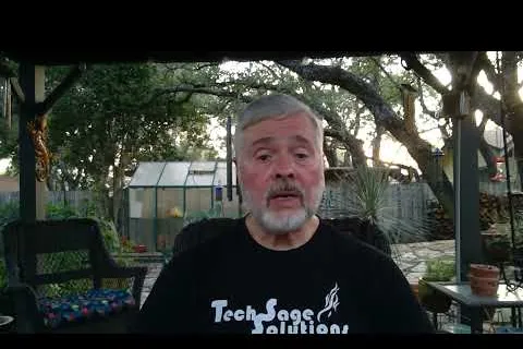 Mature man with gray hair and beard speaking outdoors, wearing a Tech Sage Solutions shirt under leafy trees.