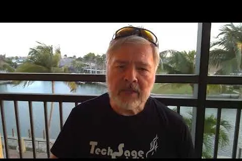 Older man with gray hair and beard wearing TechSage shirt and sunglasses on head on balcony by water and palm trees.