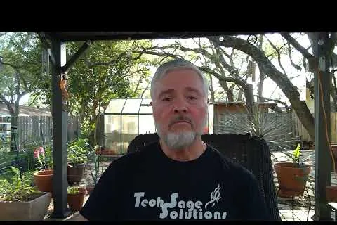 Older man with gray beard wearing Tech Sage Solutions shirt sitting outdoors in garden patio surrounded by plants and trees