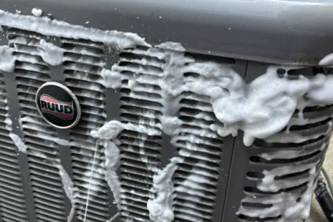 Foamy soap covers a RUUD air conditioning unit during cleaning, showcasing maintenance process outdoors.