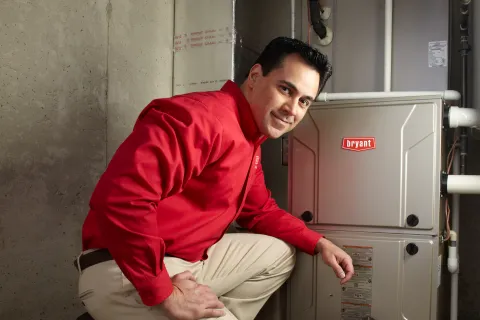 Technician in red shirt kneeling next to Bryant HVAC unit in a basement with tools.