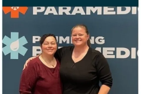Two smiling women standing in front of a Plumbing Paramedics branded wall with logo and text.