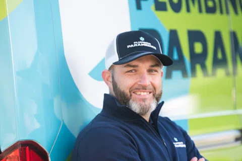 Smiling plumber in uniform and cap standing with arms crossed by a blue and green Plumbing Paramedics van.