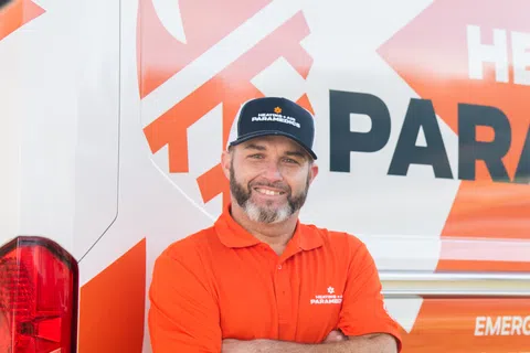 Man in bright orange shirt and black cap stands with arms crossed beside branded service van.